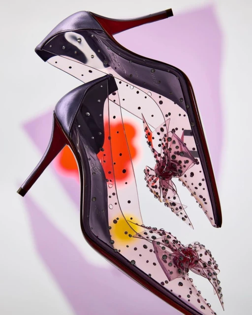 High-heeled shoes with a pointed toe, transparent material, and polka dot design, complemented by decorative bows against a colorful background.