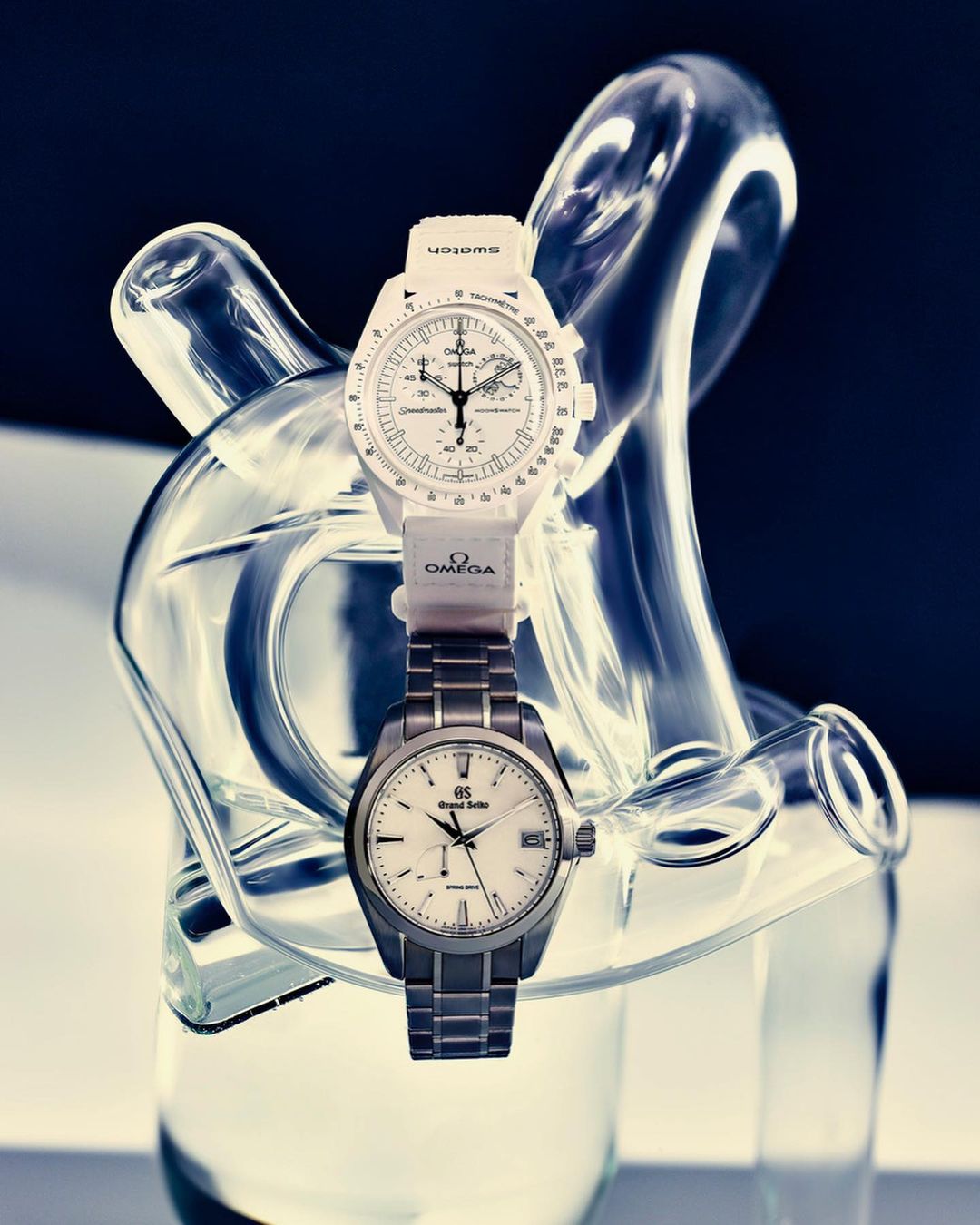 Two wrist watches, one white, one steel, presented on a clear decorative glass stand against a dark background. High-key studio lighting.