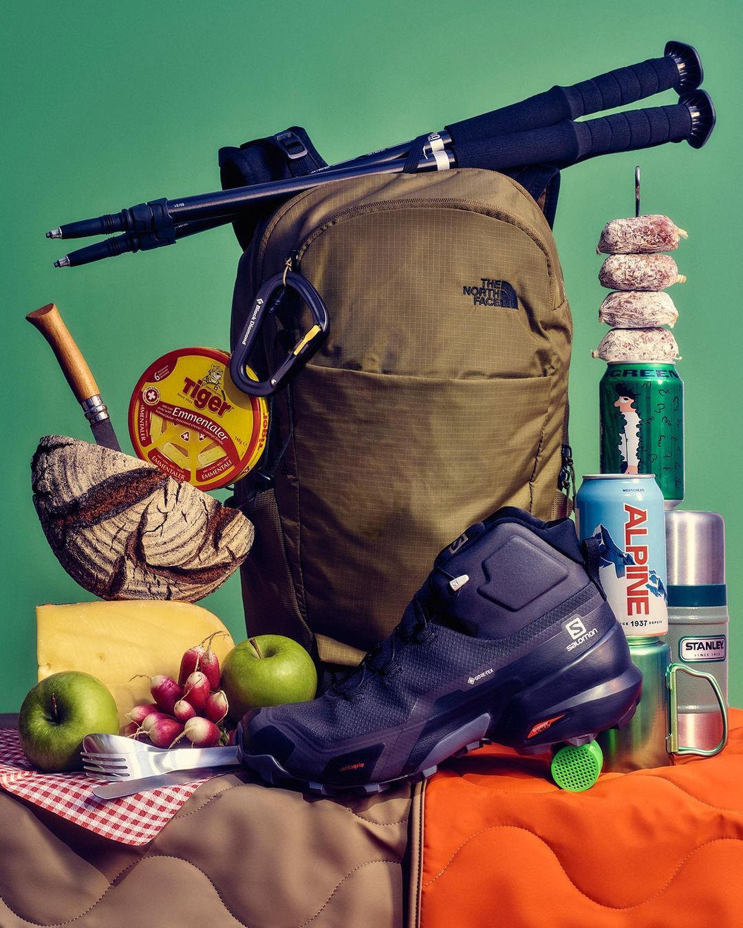 Still life of hiking gear including a backpack, hiking boots, walking sticks, and snacks against a green background.