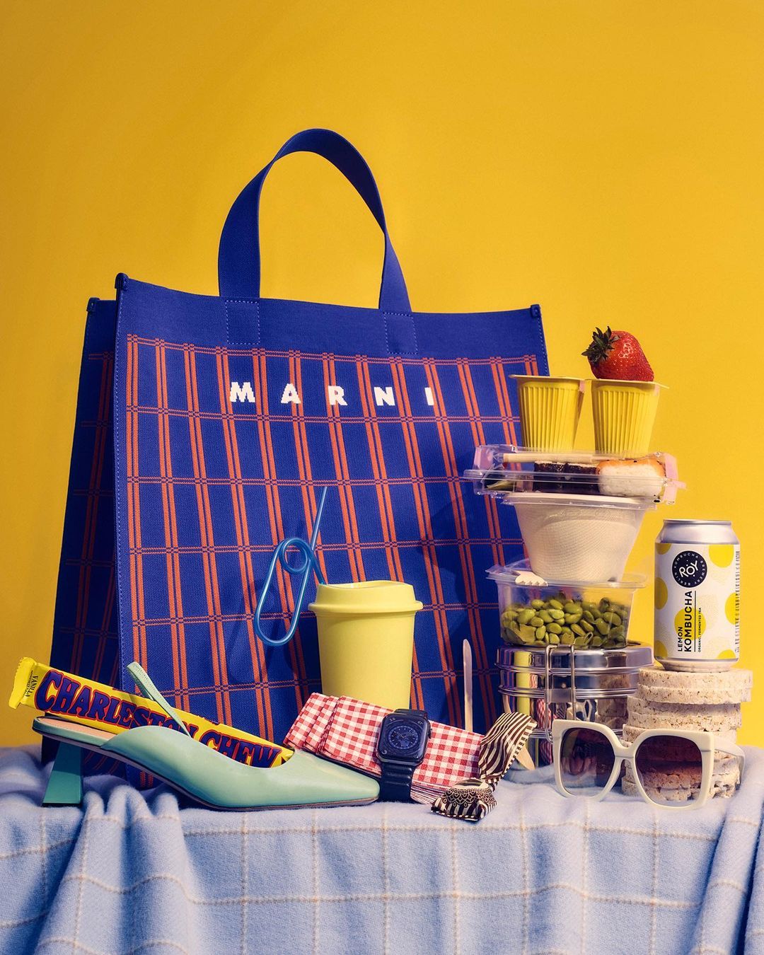 Still life of a patterned Marni tote bag with accessories, food containers, and sunglasses against a solid yellow background.