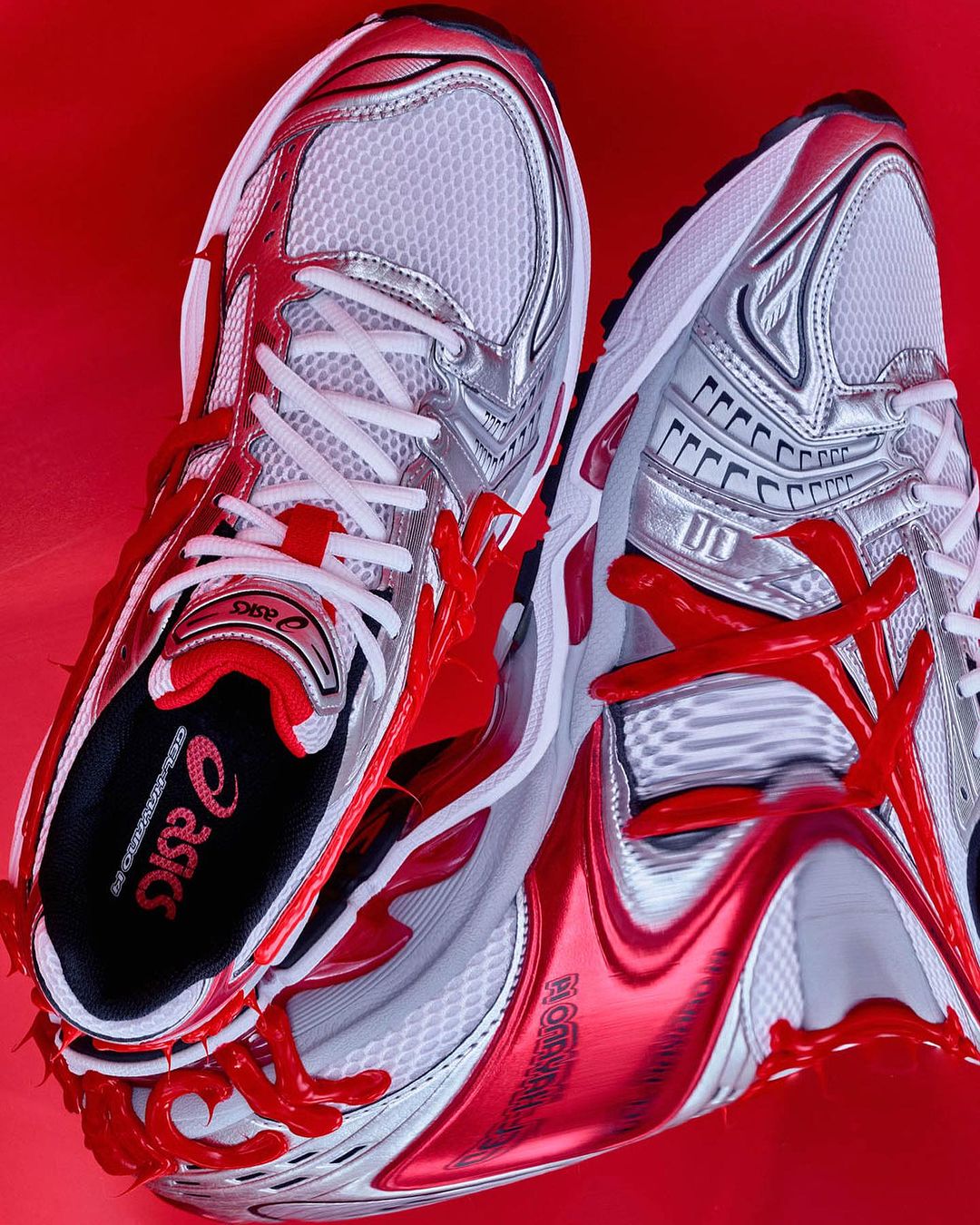 Two Asics running shoes are arranged on a reflective red surface, showcasing the brand logo, white mesh, silver accents, and red laces.