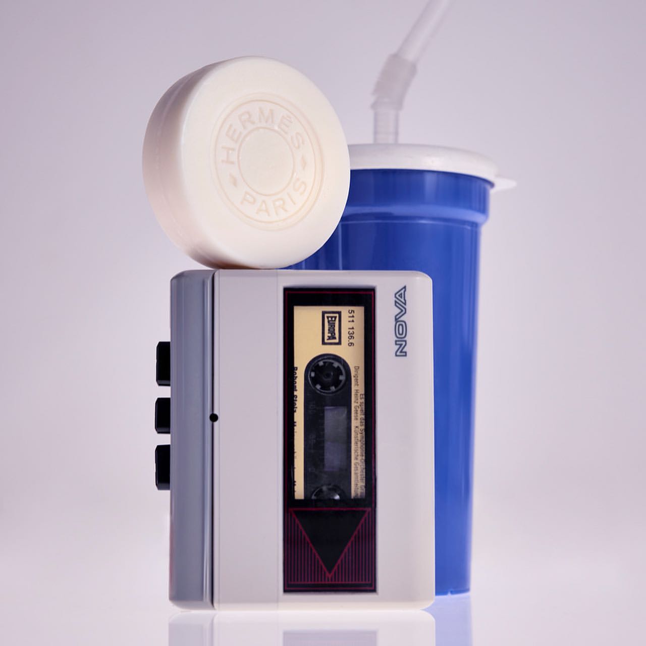 Retro-style product shot featuring a vintage cassette player, a round soap with an Hermes logo, and a blue plastic cup with a straw.