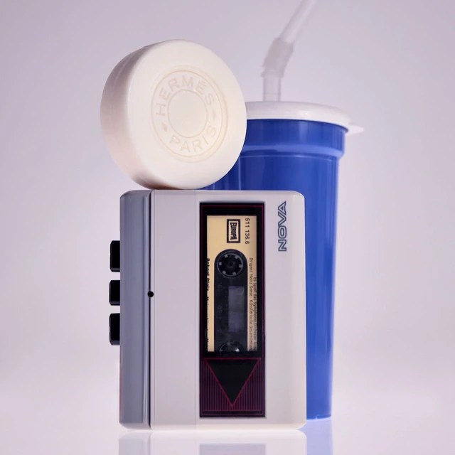 Retro-style product shot featuring a vintage cassette player, a round soap with an Hermes logo, and a blue plastic cup with a straw.