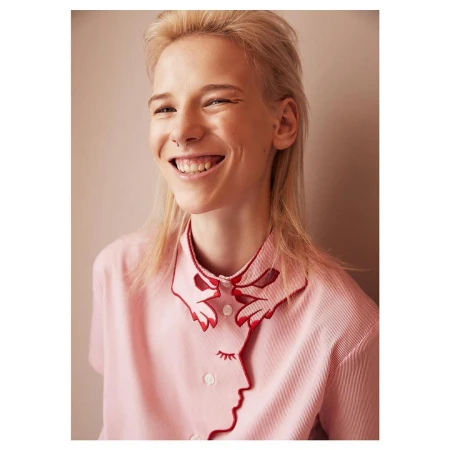 Portrait of a smiling blond person wearing a light pink shirt with distinctive red-trimmed embroidered collar details.