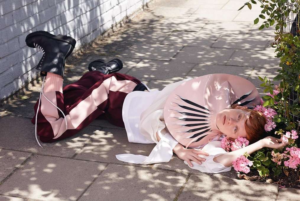A young adult female fashion model lies on pavement next to flowers wearing an avant-garde pink collar and black boots in outdoor lighting.