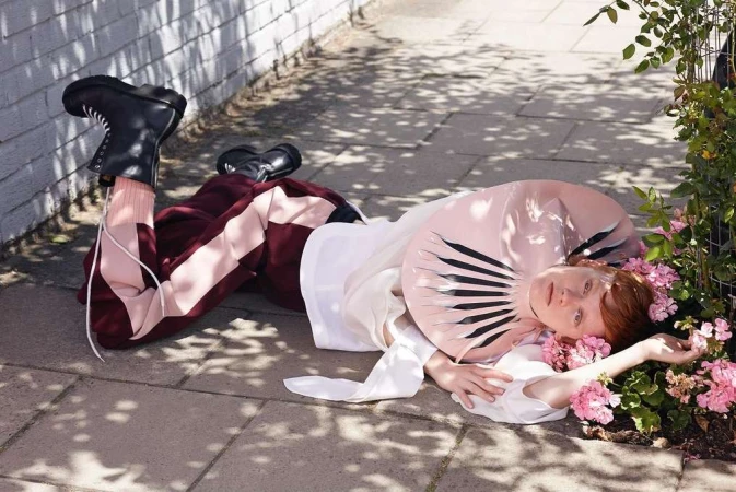 A young adult female fashion model lies on pavement next to flowers wearing an avant-garde pink collar and black boots in outdoor lighting.