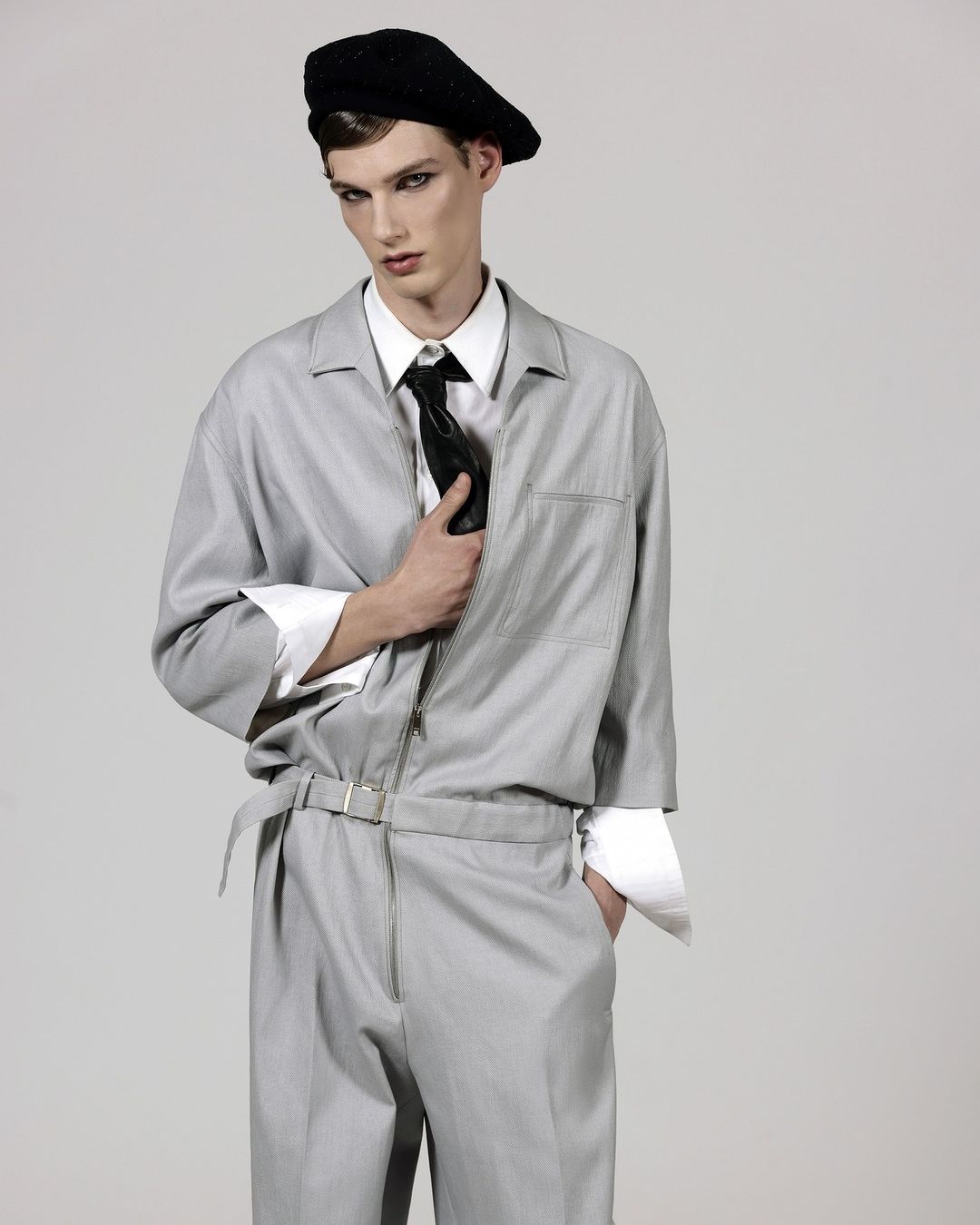 A young man poses in a light gray jumpsuit with a black beret and tie against a neutral background in a studio fashion shot.
