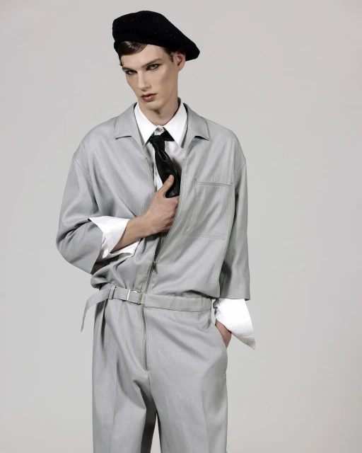 A young man poses in a light gray jumpsuit with a black beret and tie against a neutral background in a studio fashion shot.