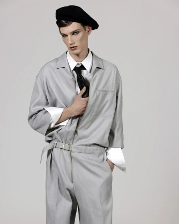 A young man poses in a light gray jumpsuit with a black beret and tie against a neutral background in a studio fashion shot.