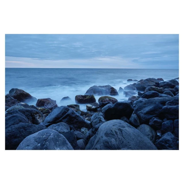 Seascape featuring dark rocks along the coastline, with the ocean water blurring due to long exposure, and a cloudy blue sky overhead.