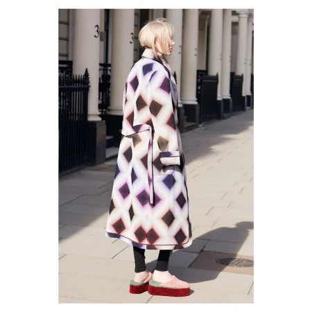 Blonde woman poses in a patterned overcoat with matching platform shoes on a sunny city street with white buildings and black iron fences.