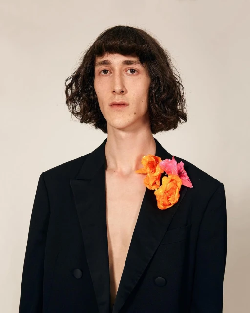 A person in a black blazer stands against a neutral backdrop, with orange and pink flowers pinned to the blazer's lapel.