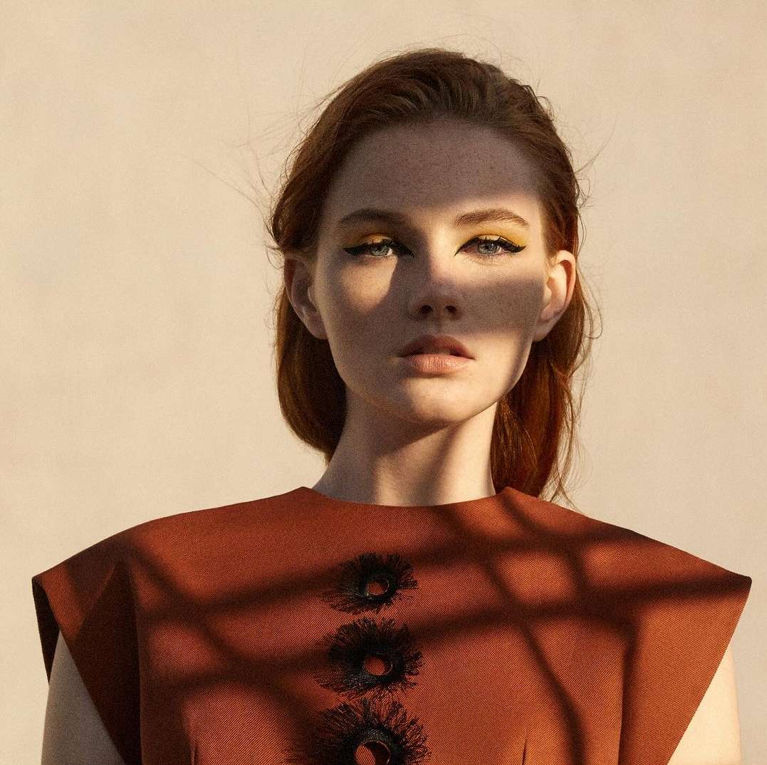 Close-up fashion portrait of a red-haired woman with yellow eyeshadow and brown dress, standing in front of a neutral background.