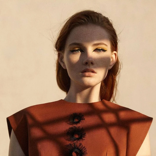 Close-up fashion portrait of a red-haired woman with yellow eyeshadow and brown dress, standing in front of a neutral background.