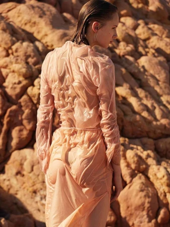 Model with wet hair wearing a sheer peach dress standing against a rocky textured background in Paris.