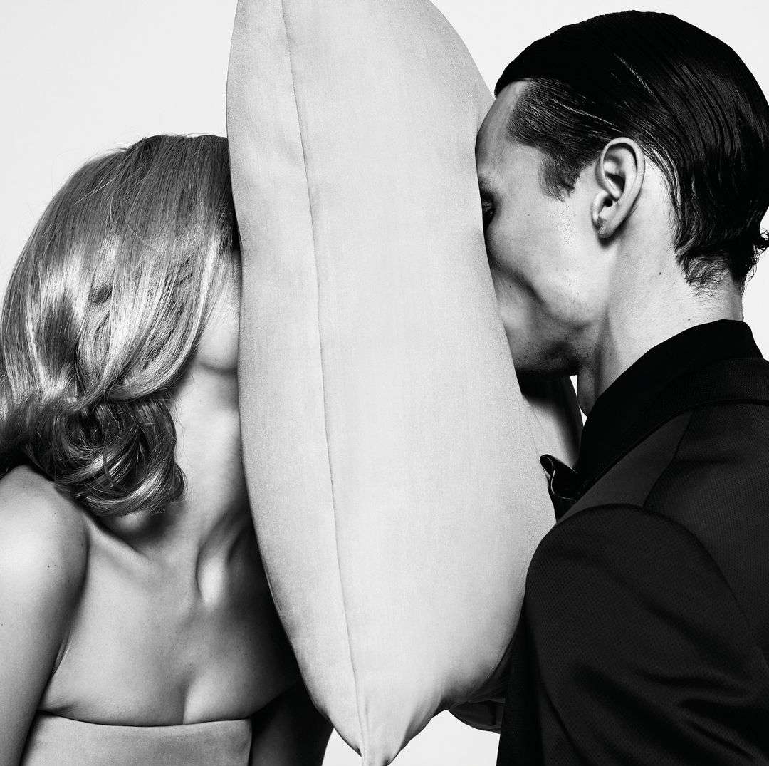 Black and white portrait of a blonde woman and dark-haired man, faces pressed against opposite sides of a light-colored square pillow.
