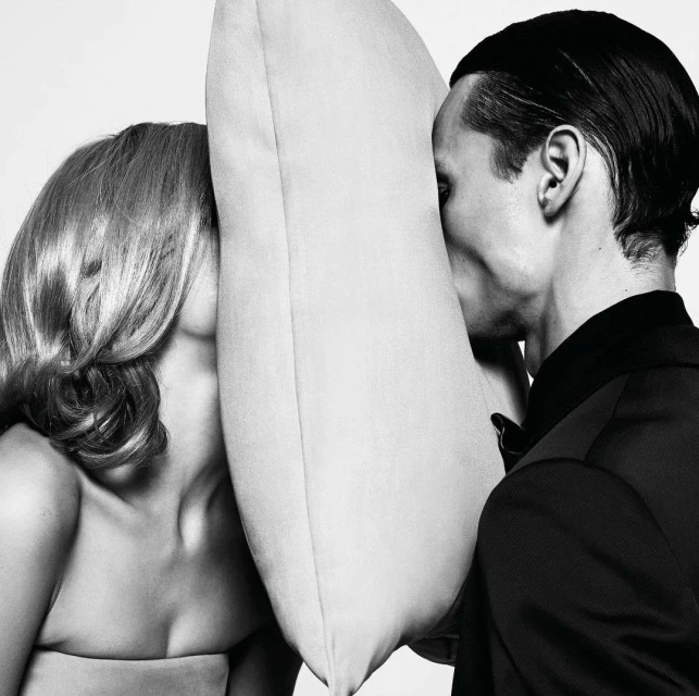 Black and white portrait of a blonde woman and dark-haired man, faces pressed against opposite sides of a light-colored square pillow.
