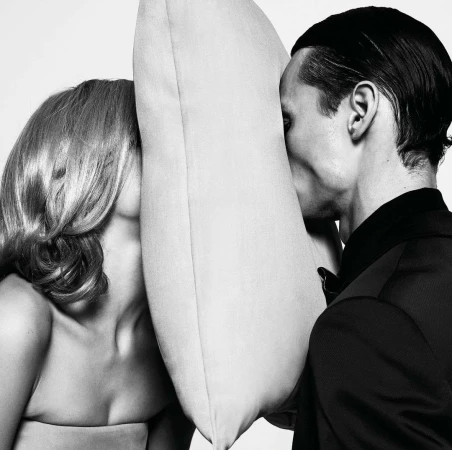 Black and white portrait of a blonde woman and dark-haired man, faces pressed against opposite sides of a light-colored square pillow.