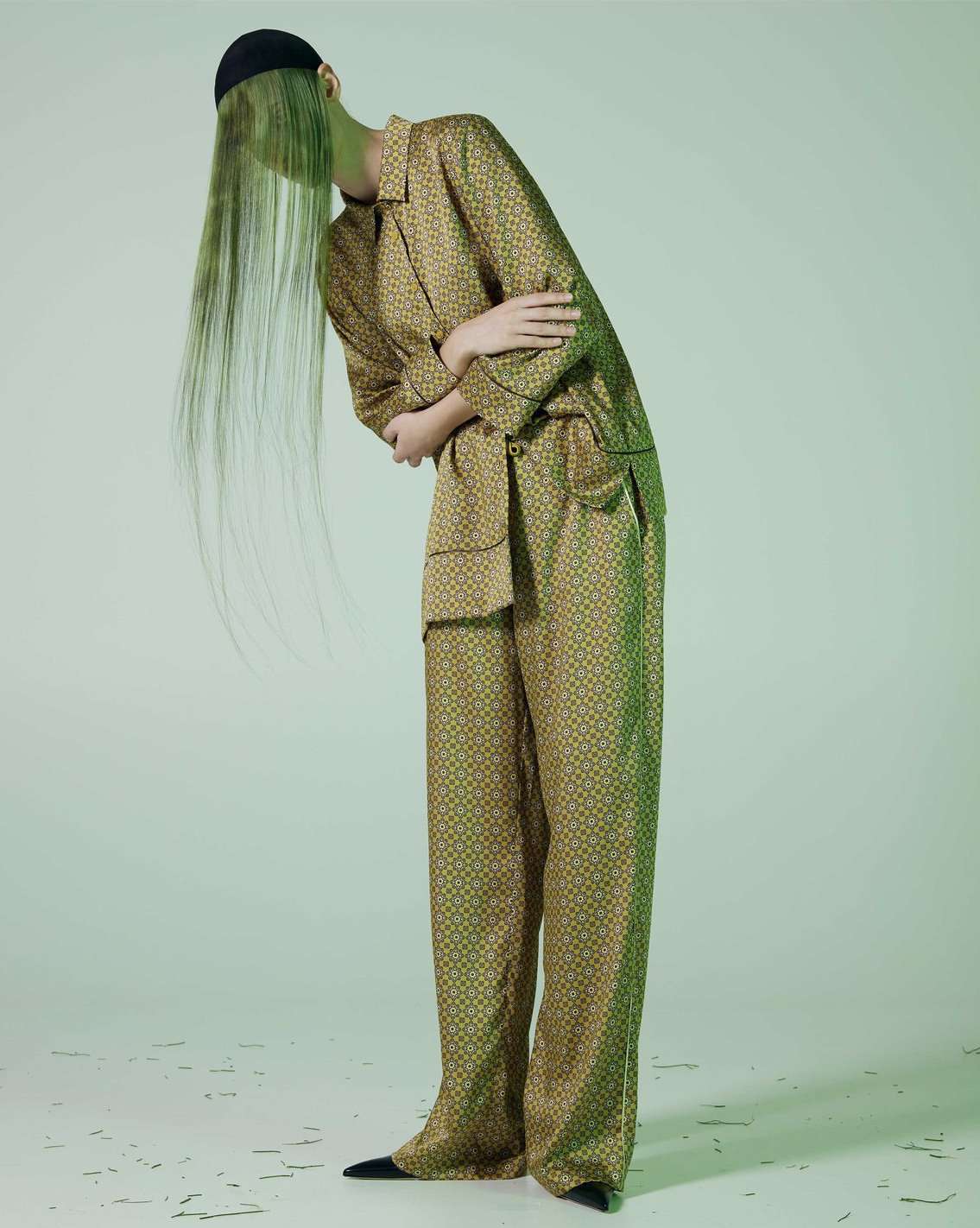 Fashion shot of a model wearing a patterned, tan pajama suit with green hair extensions and a black cap, against a gradient green backdrop.
