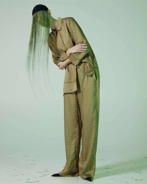 Fashion shot of a model wearing a patterned, tan pajama suit with green hair extensions and a black cap, against a gradient green backdrop.