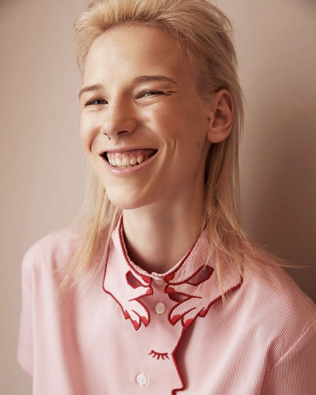 Smiling young adult female with blonde hair, wearing a pink shirt with red trim, and a nose ring, looking directly at the camera.