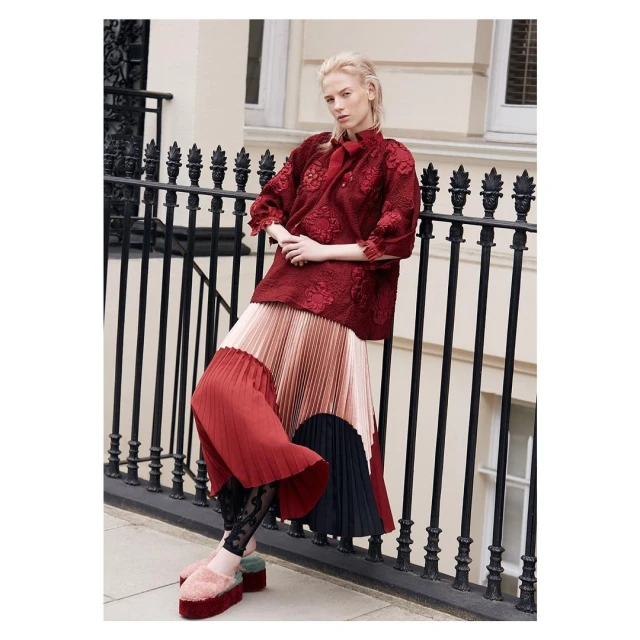 Fashionable woman in a red floral top, pleated skirt, and platform shoes poses against a London building and wrought-iron railing.