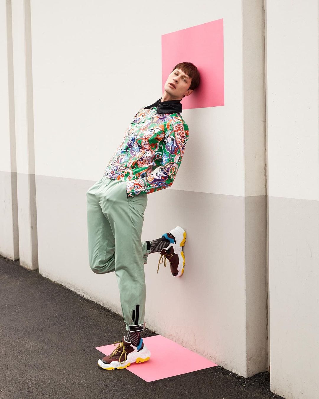 Stylish male model in patterned shirt and green pants leans against a wall with pink squares, showcasing contemporary fashion.
