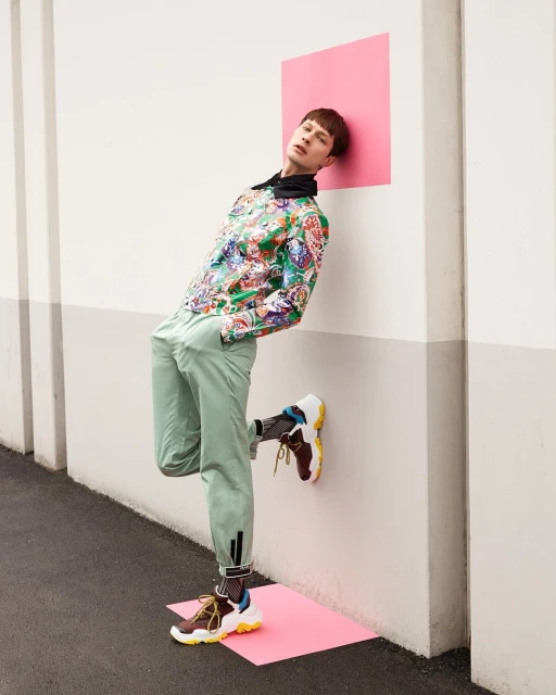 Stylish male model in patterned shirt and green pants leans against a wall with pink squares, showcasing contemporary fashion.