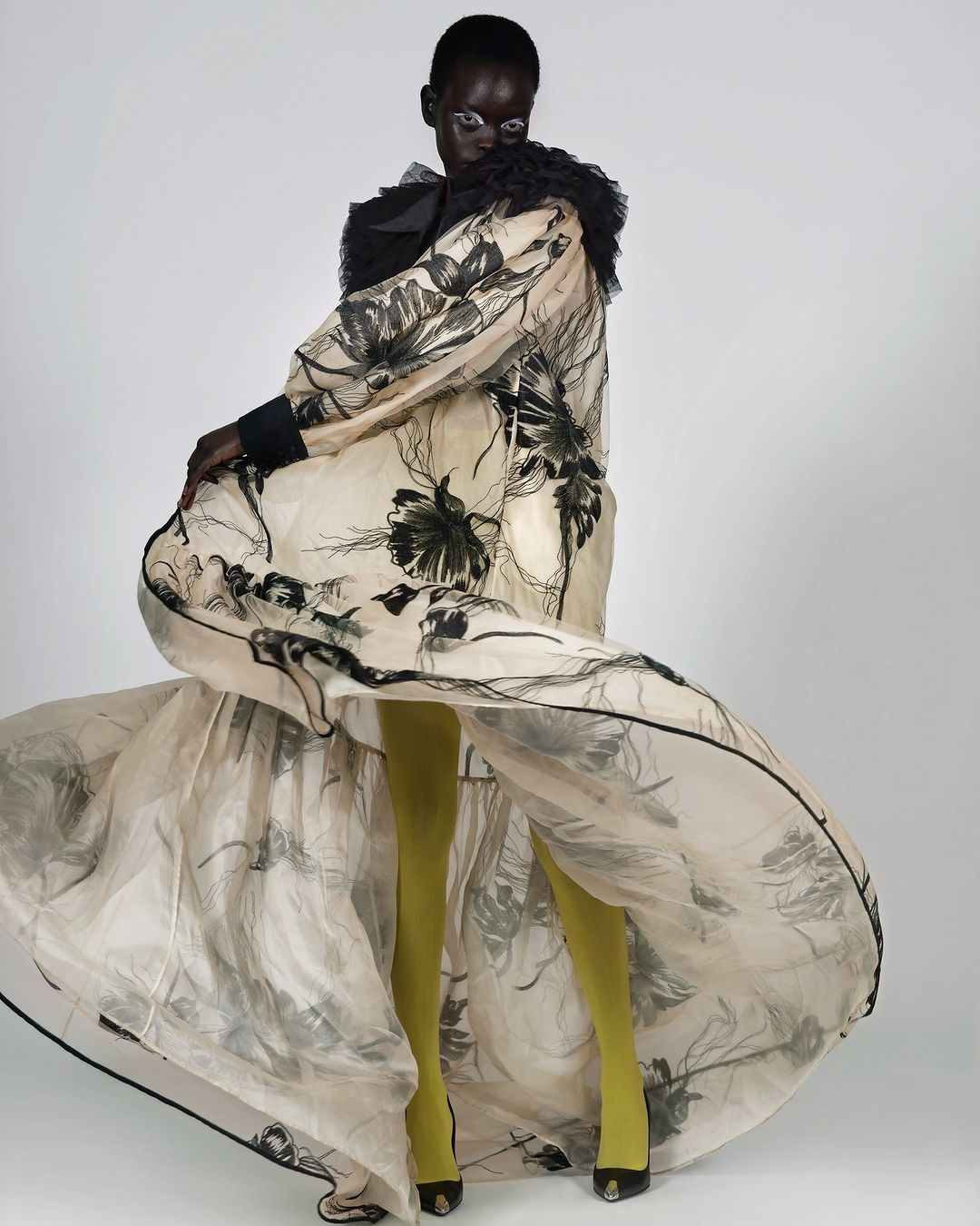 Woman in flowing floral gown with sheer fabric, olive tights, and heels, captured in a fashion forward studio shot against a white backdrop.