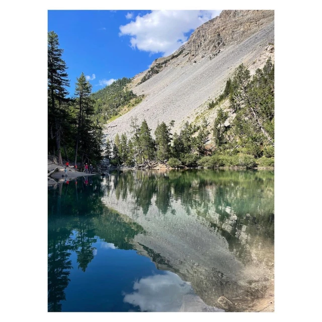 Scenic view of a lake reflecting a rocky mountain and trees under a blue sky with clouds, with several people along the shoreline.