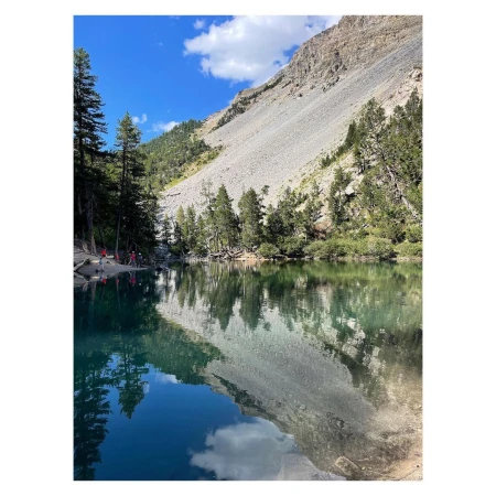 Scenic view of a lake reflecting a rocky mountain and trees under a blue sky with clouds, with several people along the shoreline.