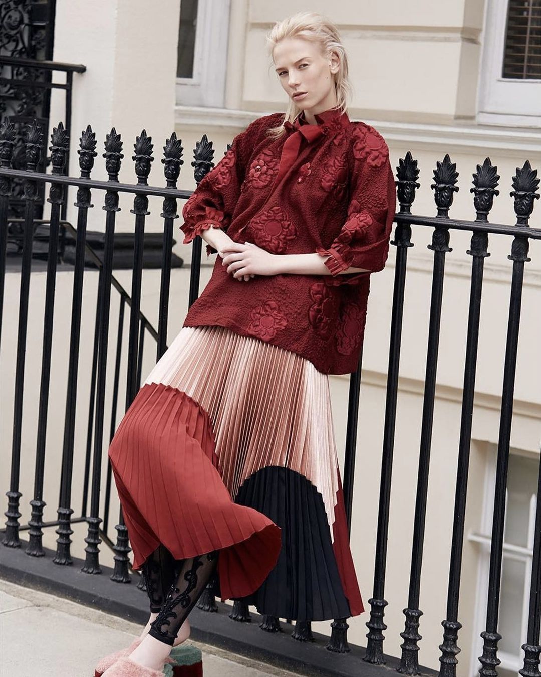 Fashion editorial image: a female model in a red top and multicolored pleated skirt leans against a black iron fence outside.