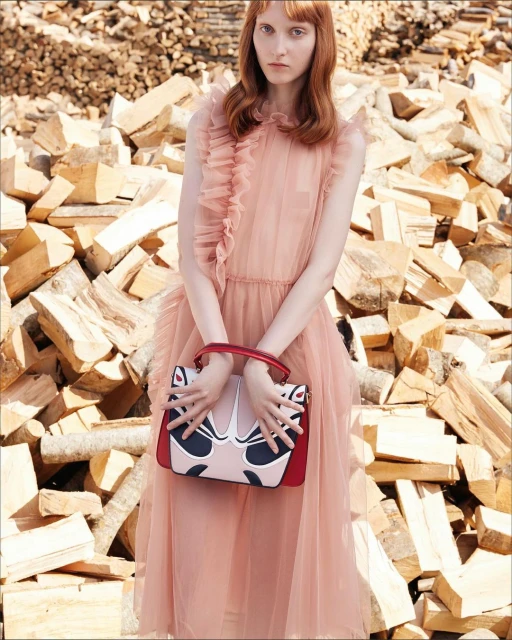 A woman in a pink dress poses in front of a wood pile, holding a colorful bag. The image highlights fashion, beauty, and editorial style.