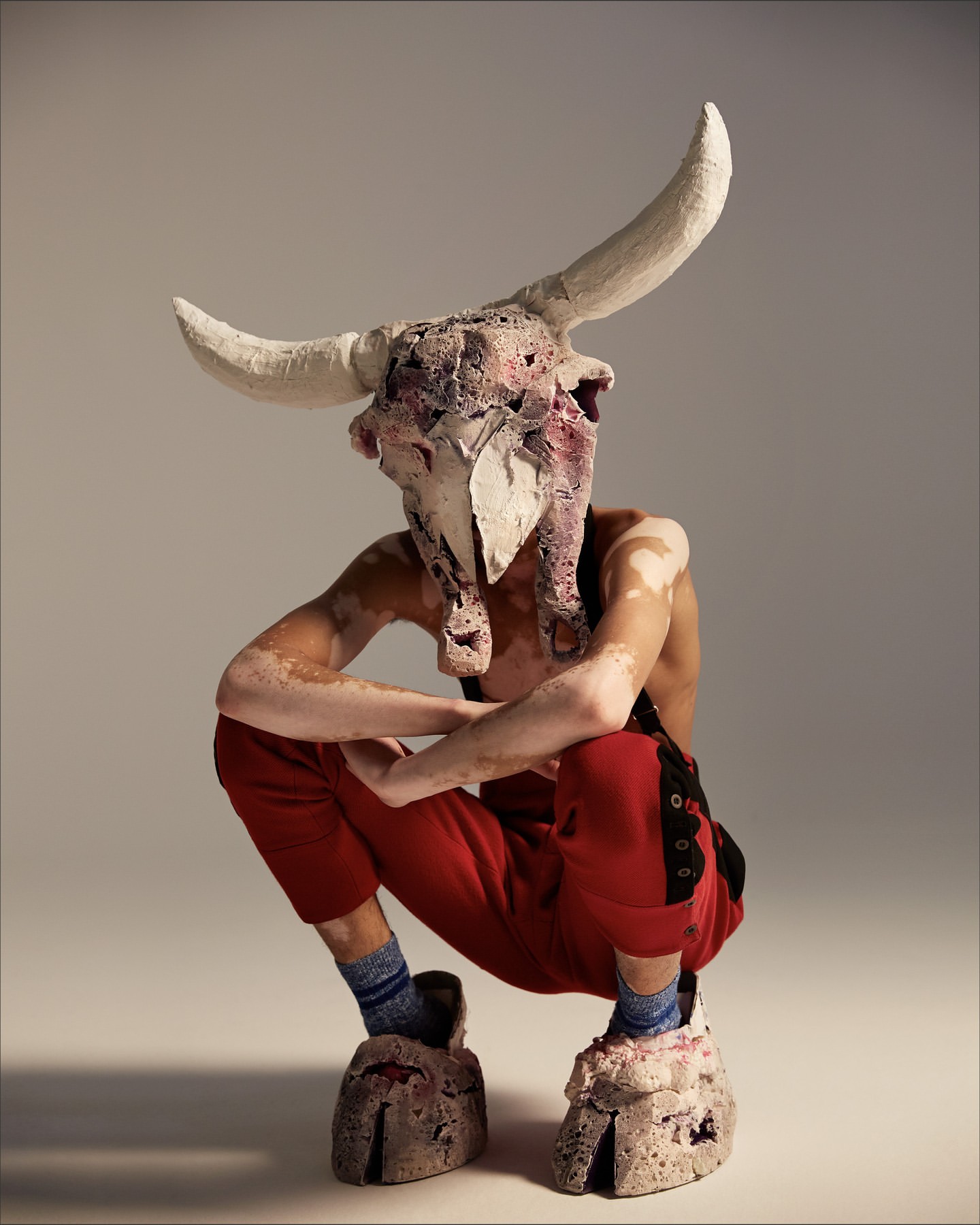 Adult human crouches in a bull skull mask, wearing red pants and oversized hoof shoes in front of a studio backdrop.