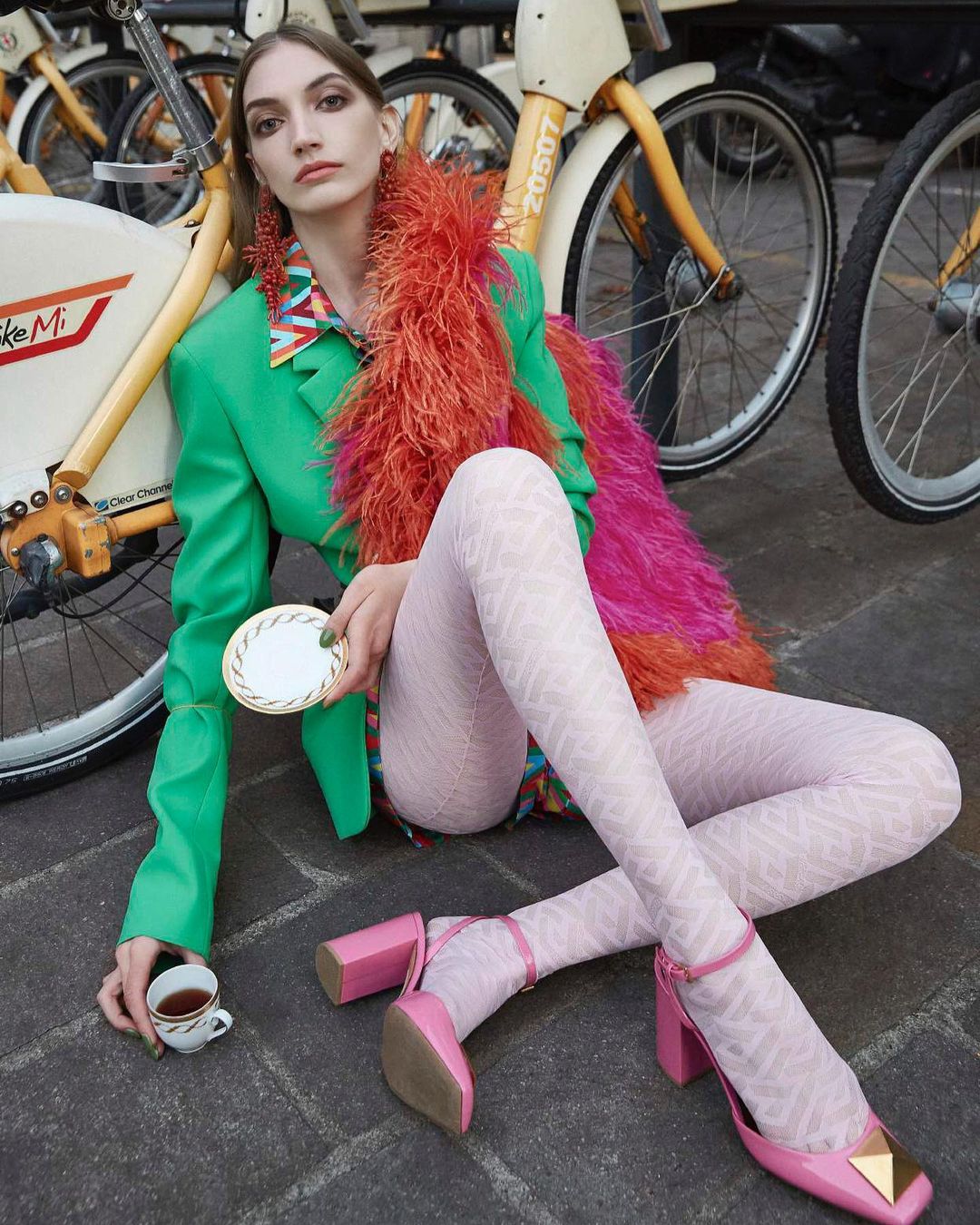 Fashionable woman poses with a teacup and bikes in an outdoor setting, wearing pink patterned tights, orange boa, green jacket, and pink shoes.
