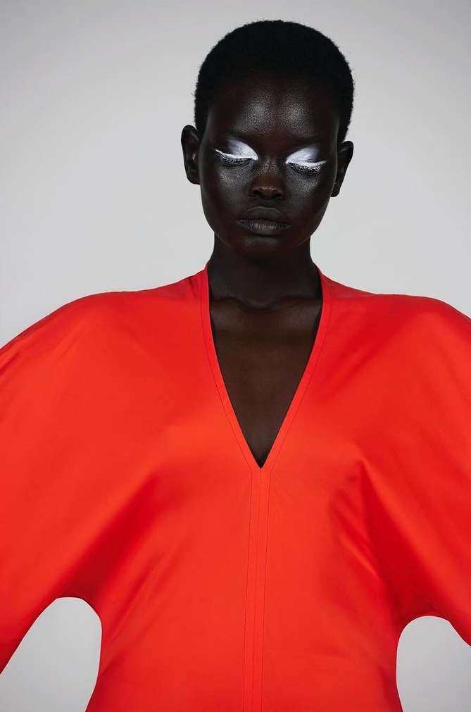 Close-up of a stylish, dark-skinned woman in a vivid red dress with creative white eye makeup, against a neutral background, fashion.