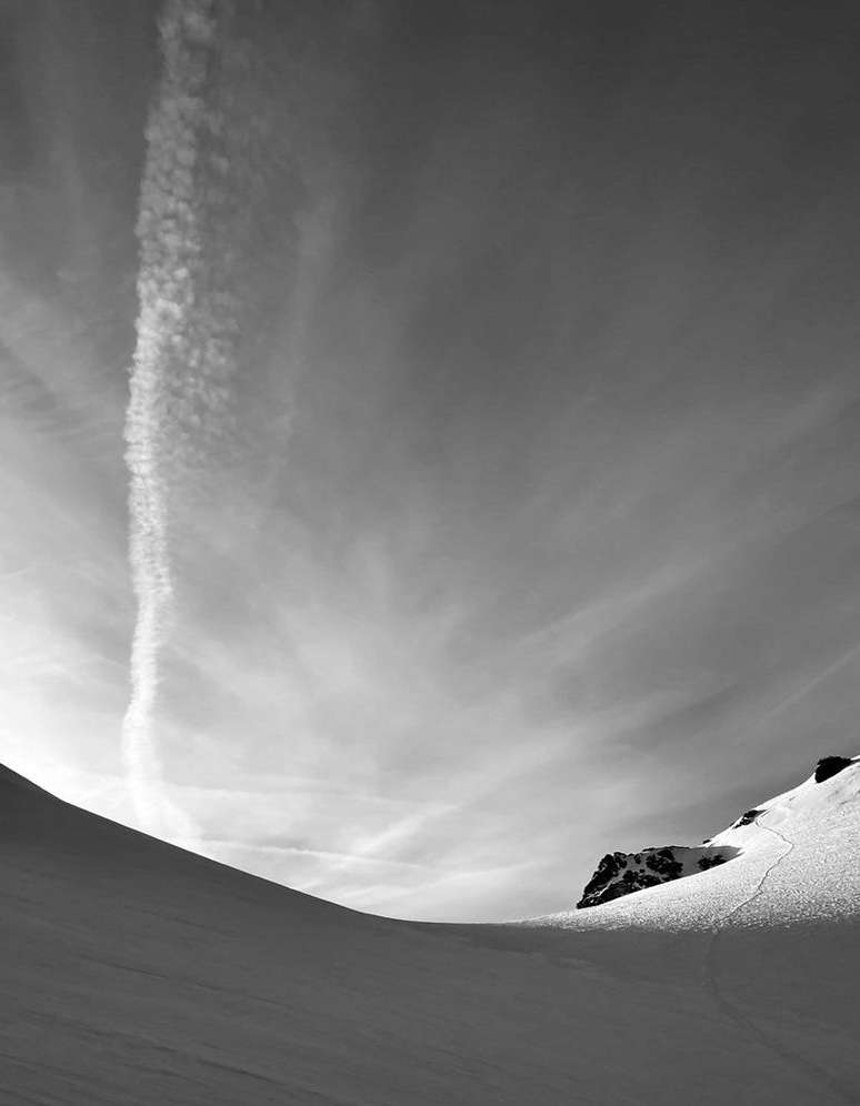 Monochrome landscape featuring snow-covered terrain under a cloudy sky with a contrail, capturing a serene and minimalist outdoor scene.