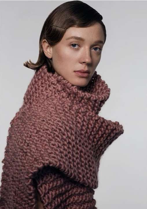 Close-up of a woman with short wavy brown hair and fair skin, wearing a chunky knit scarf in a neutral setting.