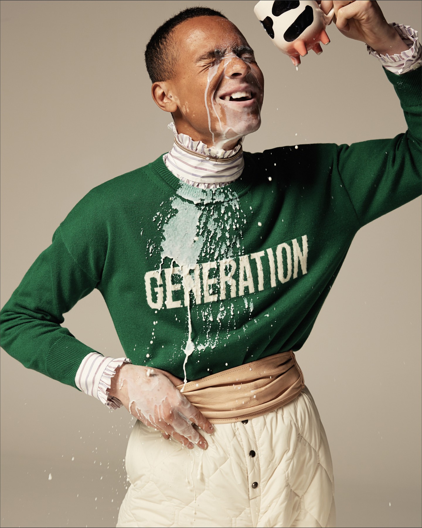 Adult male model in a studio with milk splashing on his face from a cow-shaped mug, wearing a green sweater and quilted pants.