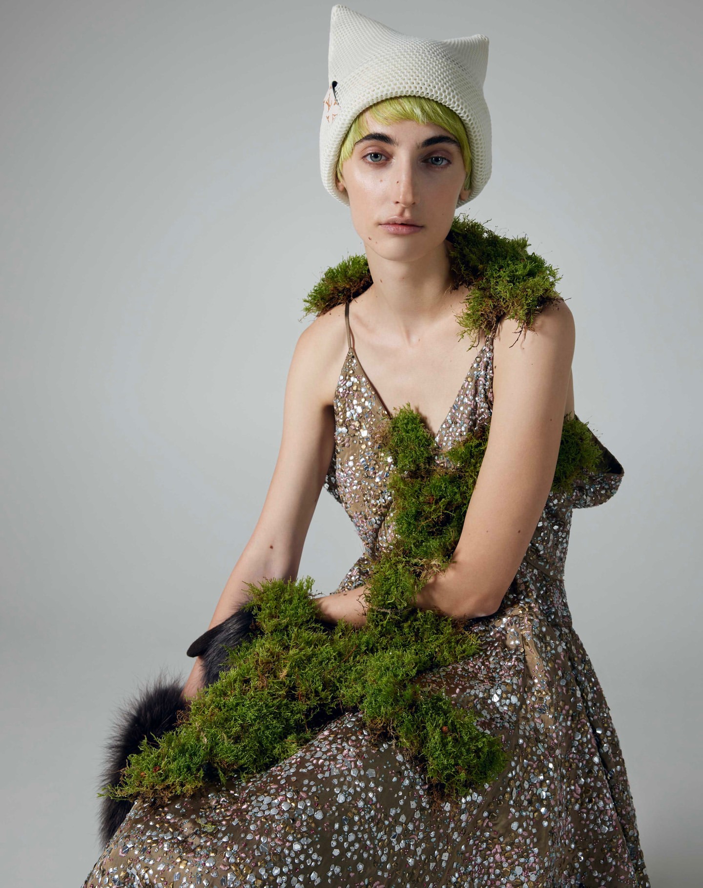 Woman in a sequined dress and a cat-eared hat, adorned with moss, poses in a studio with soft lighting against a gray backdrop.