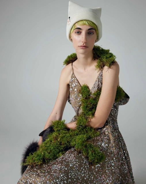 Woman in a sequined dress and a cat-eared hat, adorned with moss, poses in a studio with soft lighting against a gray backdrop.