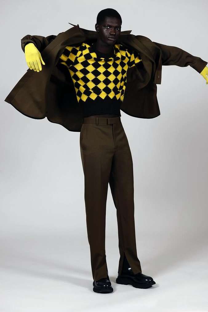 Dark-skinned male model wears a dark brown suit and checkerboard sweater with yellow gloves, standing in a studio setting against a white backdrop.