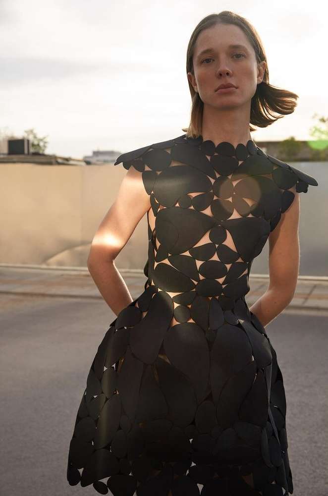 Woman in a short black dress with geometric cutouts, posing on a street with a concrete wall in the background, illuminated by natural light.