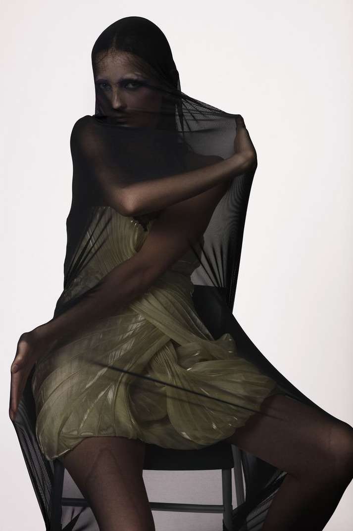 A woman draped in sheer black fabric poses in a studio setting, wearing a light gold dress; the image is a fashion studio portrait.