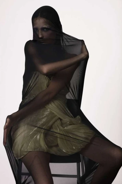 A woman draped in sheer black fabric poses in a studio setting, wearing a light gold dress; the image is a fashion studio portrait.