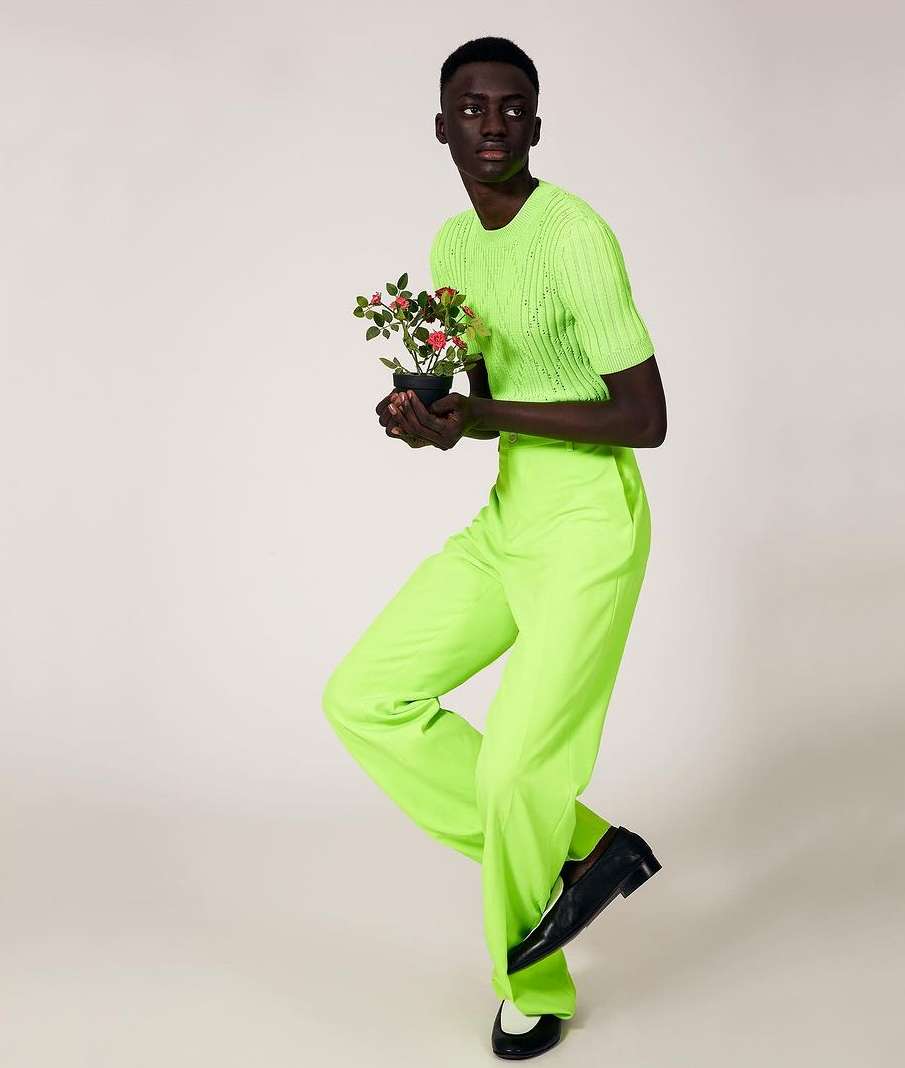 Dark-skinned man in neon green outfit posing with a potted plant on a light background in a fashion shot. Full body view.
