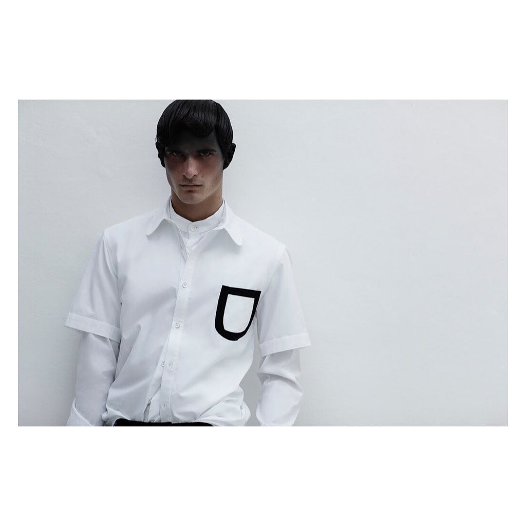 Fashion shot of a male model in a white button-down shirt with a bold black pocket, against a stark white background with diffused lighting.