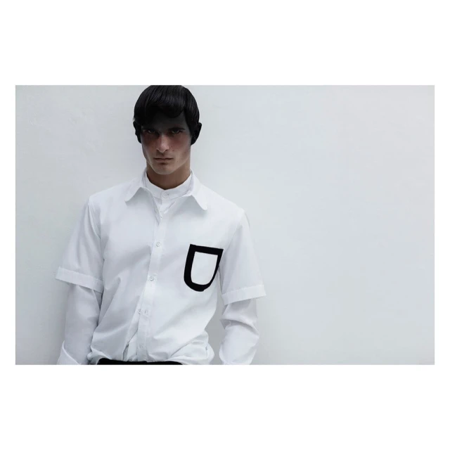 Fashion shot of a male model in a white button-down shirt with a bold black pocket, against a stark white background with diffused lighting.