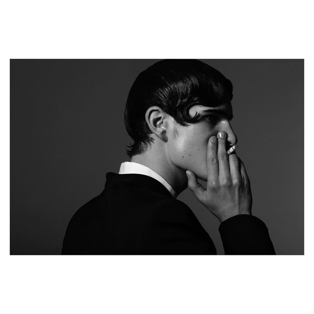 Monochrome portrait of a man in a suit, with slicked-back hair, holding a cigarette and his hand to his face against a grey backdrop.