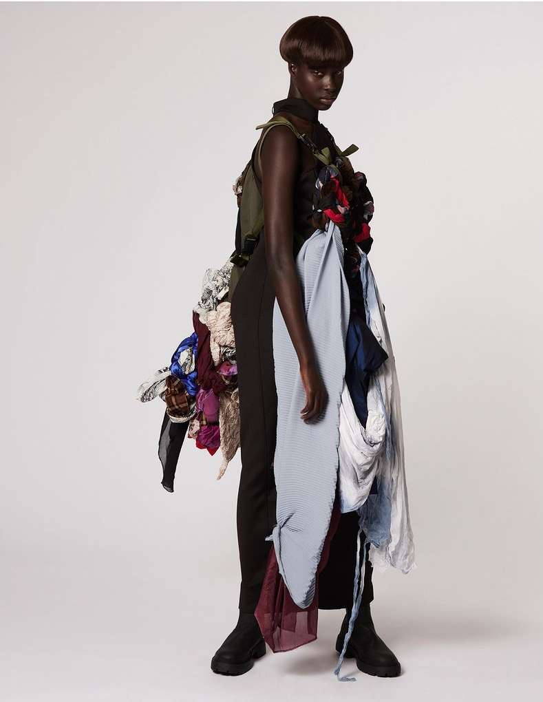 Model showcases avant-garde fashion with layered fabrics and textures, set against a clean white backdrop in a studio setting.